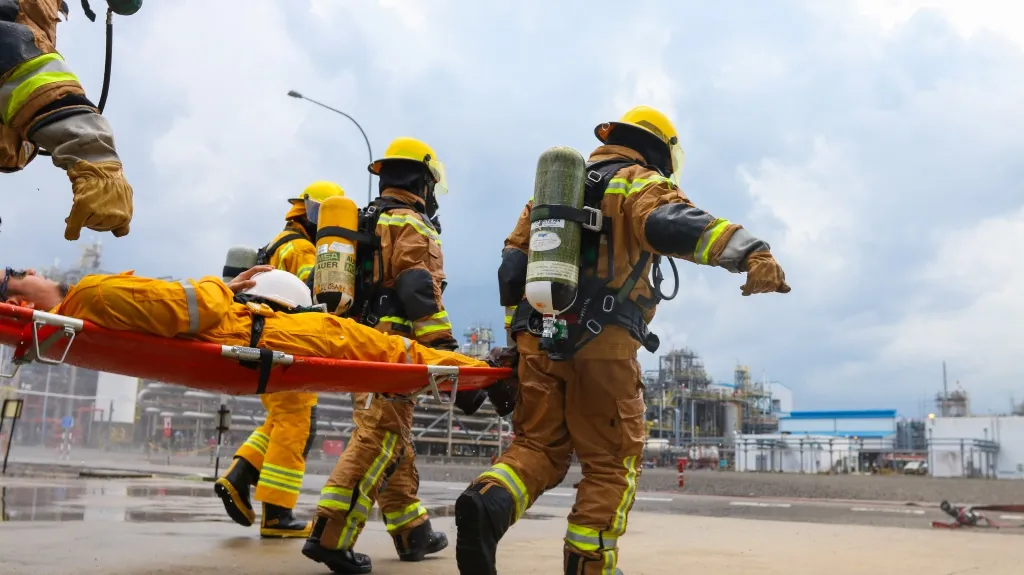 Firefighters in full gear carrying an injured person on a stretcher at an industrial site.