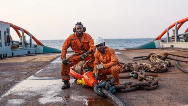 Two male workers in orange coveralls working with large chains and equipment on a ship deck by the sea.
