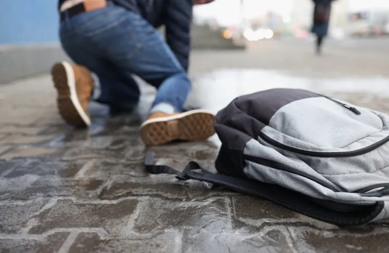Gray and black backpack on wet pavement with a person kneeling in jeans and brown shoes.