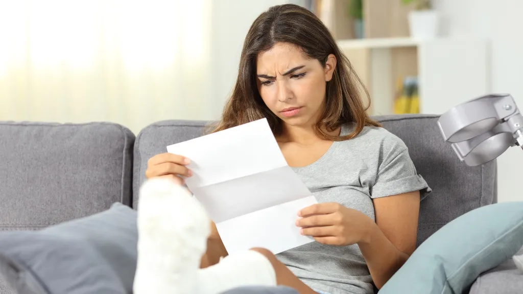 Young woman with a leg cast sitting on gray couch, reading a letter with a concerned expression.