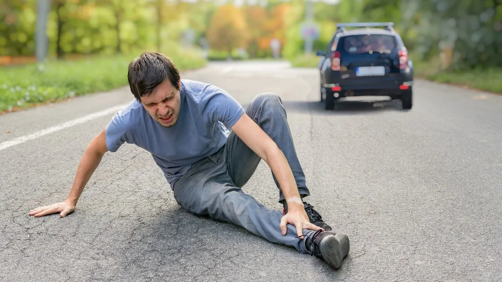 Man in casual clothing sitting on a road, holding his ankle in pain with a black car in the background.