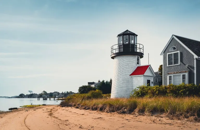 White cylindrical lighthouse with black top and red-roofed building beside a sandy beach.